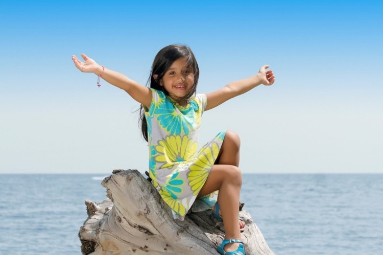 Young girl with long brown hair wearing a girls Hawaiian dress with big flowers on it, sitting on driftwood with arms outreached and calm ocean waters in the background.