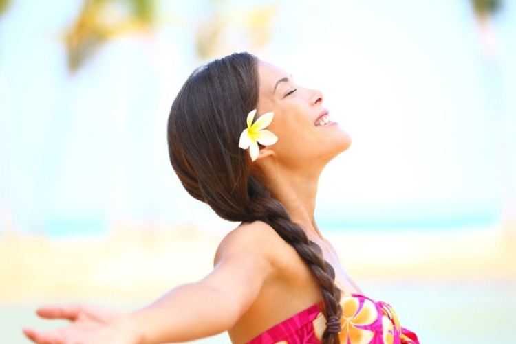 Pretty young woman with a white and yellow hibiscus flower in her hair, wearing a classic floral Hawaiian dress, looking to the sky with eyes closed, smiling, and arms outstretched.